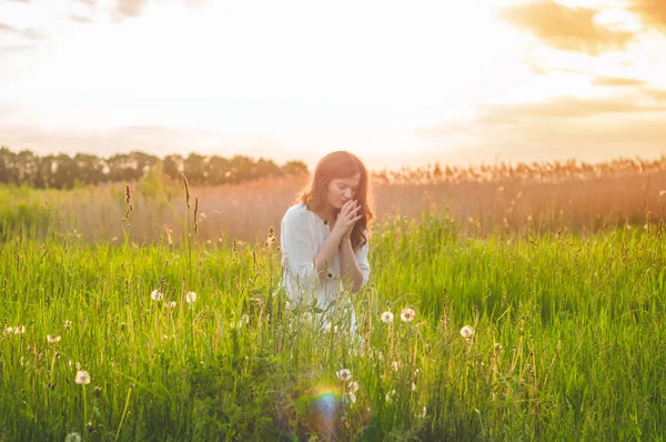 Girl closed her eyes, praying in a field during beautiful sunset. Hands folded in prayer concept for faith