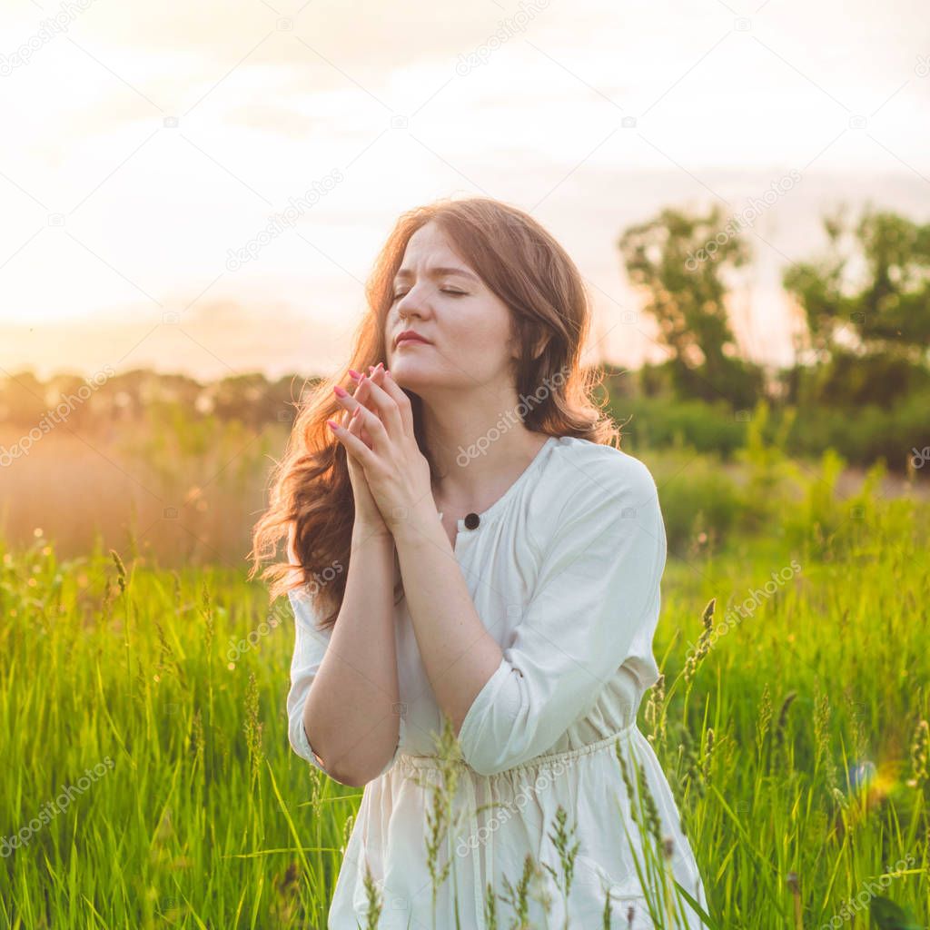 Girl closed her eyes, praying in a field during beautiful sunset. Hands folded in prayer concept for faith