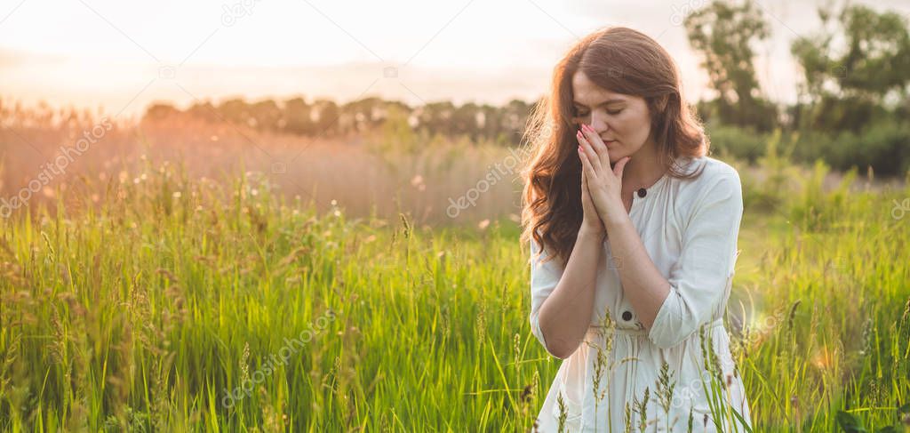 Girl closed her eyes, praying in a field during beautiful sunset. Hands folded in prayer concept for faith