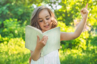 Şirin, çalışkan bir çocuk elinde bir kitap ve açık havada bir çantayla ayakta duruyor. Kitabı okuyan küçük kız.