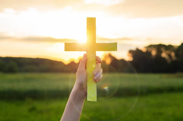 Gadis remaja memegang salib di tangan selama matahari terbenam yang indah. Tangan dilipat dalam konsep doa untuk iman — Stok Foto
