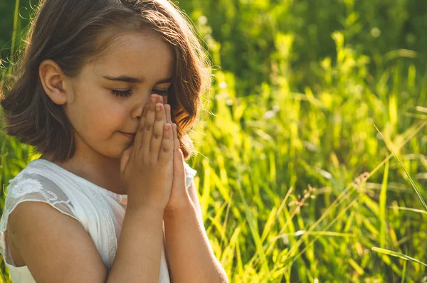Gadis kecil menutup matanya, berdoa di lapangan selama matahari terbenam yang indah. Tangan dilipat dalam konsep doa untuk iman — Stok Foto