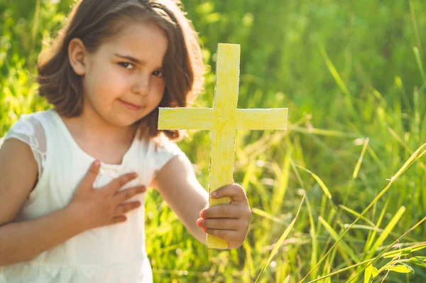 Niña sosteniendo la cruz en la mano durante el hermoso atardecer. Manos dobladas en concepto de oración por la fe —  Fotos de Stock