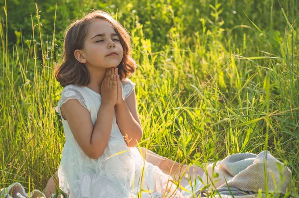 Little Girl becsukta a szemét, imádkozott a területen során gyönyörű naplemente. Az imádság koncepciójában kézzel hajtogatott kezek — Stock Fotó