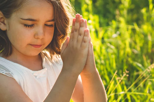 Маленька дівчинка закрила очі, молячись в полі під час прекрасного заходу сонця. Руки складені в молитві концепція віри — стокове фото