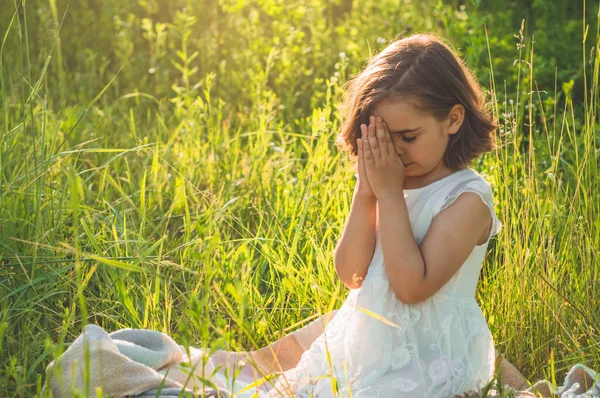 Bambina chiuse gli occhi, pregando in un campo durante il bel tramonto. Mani giunte nel concetto di preghiera per la fede Foto Stock Royalty Free