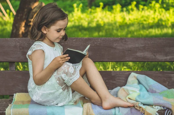 A menina tem o livro nas mãos. A ler o livro ao ar livre. A menina sentada em um banco — Fotografia de Stock