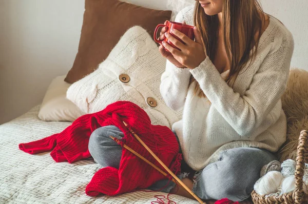 Krásná dívka si odkrájí Teplý svetr s horkou šálkem čaje na posteli. Pletení jako hobby — Stock fotografie