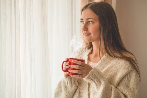 Mujer con una taza de bebida caliente junto a la ventana. Mirando la ventana y bebiendo té. Buenos días con el té. — Foto de Stock