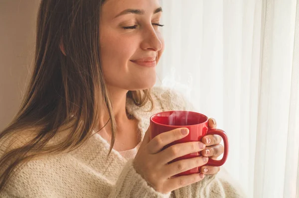 Mujer con una taza de bebida caliente junto a la ventana. Mirando la ventana y bebiendo té. Buenos días con el té. — Foto de Stock