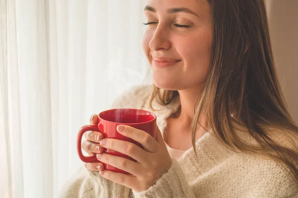 Mujer con una taza de bebida caliente junto a la ventana. Mirando la ventana y bebiendo té. Buenos días con el té. . — Foto de Stock