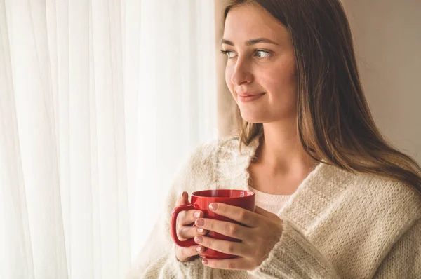 Mujer con una taza de bebida caliente junto a la ventana. Mirando la ventana y bebiendo té. Buenos días con el té. . — Foto de Stock