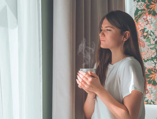 Mujer con una taza de bebida caliente junto a la ventana. Mirando la ventana y bebiendo té. Buenos días con el té. . — Foto de Stock
