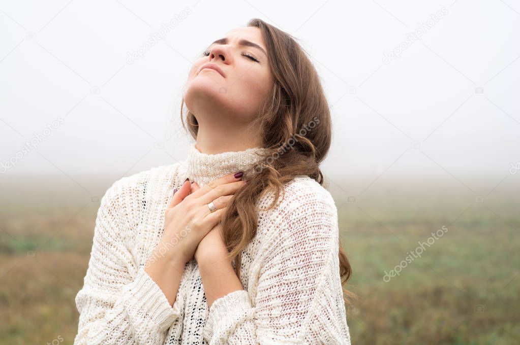 Girl closed her eyes, praying in a field during beautiful fog. Hands folded in prayer concept for faith