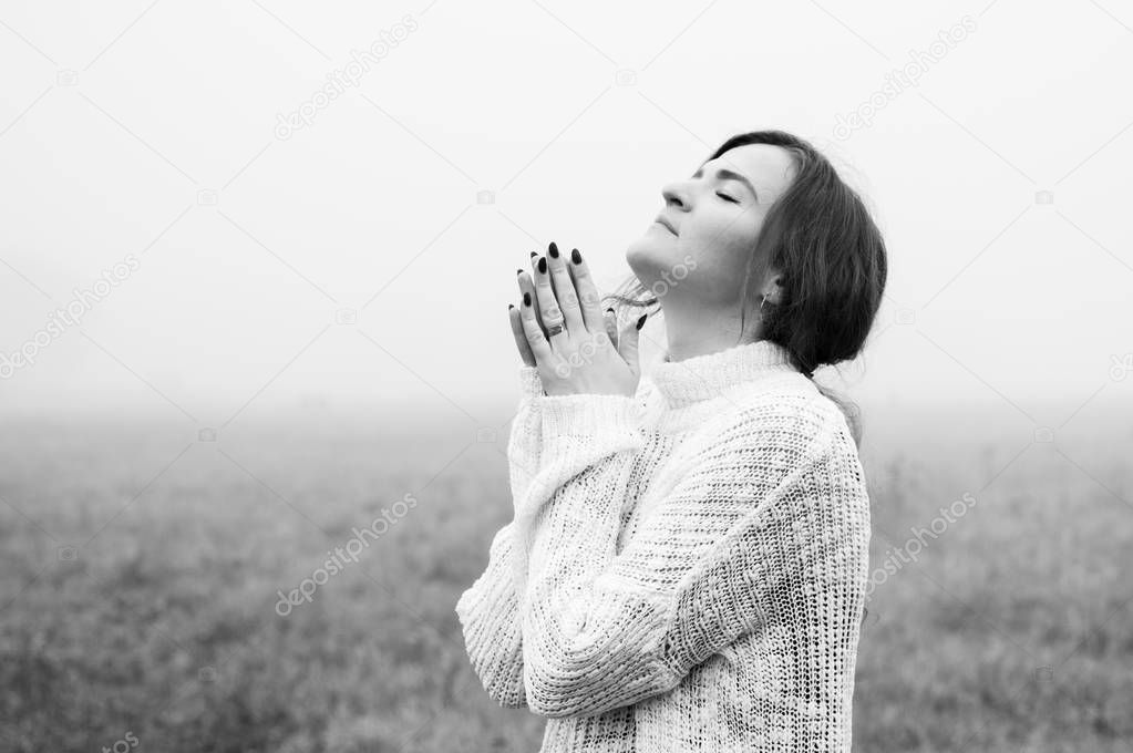 Girl closed her eyes, praying in a field during beautiful fog. Hands folded in prayer concept for faith