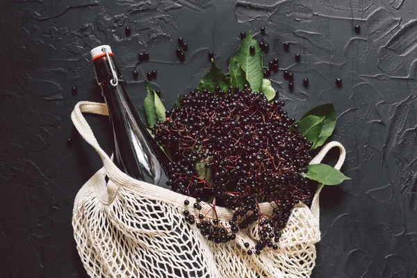 A bottle of elderberry syrup on a dark background with fresh elderberries and string bag — Stock Photo, Image