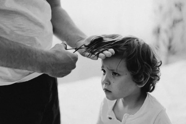 Black and white photo. Father cuts her son hair in the room. Family during quarantine, Scissors and a comb in male hands — Stock Photo, Image