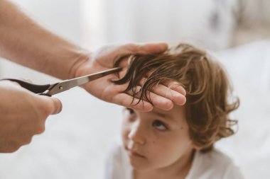 Baba odada oğlunun saçını kesiyor. Karantina sırasındaki aile, makas ve erkek ellerinde tarak.