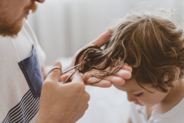 Baba odada oğlunun saçını kesiyor. Karantina sırasındaki aile, makas ve erkek ellerinde tarak.