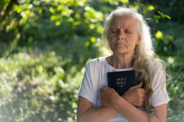 A avó de cabelos grisalhos tem a bíblia nas mãos. Lendo a Bíblia Sagrada em uma natureza. Conceito de fé, espiritualidade e religião — Fotografia de Stock