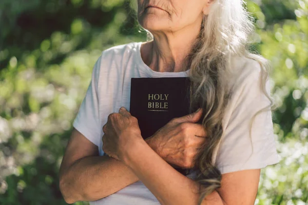 A avó de cabelos grisalhos tem a bíblia nas mãos. Lendo a Bíblia Sagrada em uma natureza. Conceito de fé, espiritualidade e religião — Fotografia de Stock