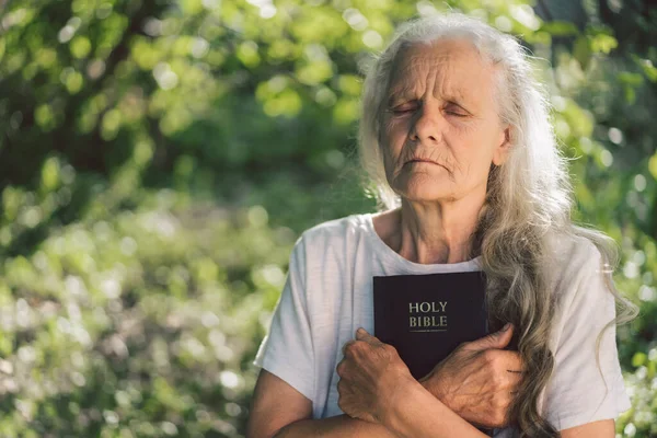 A avó de cabelos grisalhos tem a bíblia nas mãos. Lendo a Bíblia Sagrada em uma natureza. Conceito de fé, espiritualidade e religião — Fotografia de Stock