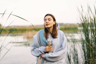 Nehir kenarında güzel bir gezgin kadın termostan sıcak bir içecek içiyor. Kadın turist nehir kenarındaki sonbahar manzarasının tadını çıkarıyor. Seyahat konsepti, rahatla..