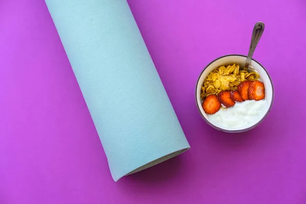 Blue mat for yoga and yoghurt with strawberries and fruits on a purple background