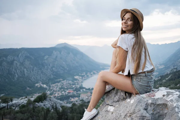 年轻微笑的旅行妇女的肖像戴着帽子坐在高高的山顶上 傍晚与日落 — 图库照片