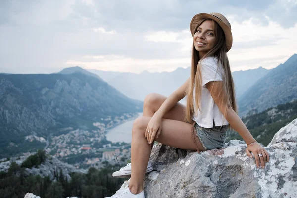 山の上に帽子座っている高身に着けている若い笑顔旅女性の肖像夕日と夜します Montenegr — ストック写真