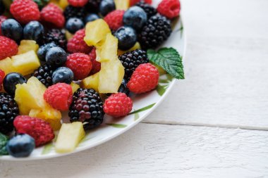 Gökkuşağı renkleri, ananas, böğürtlen, yaban mersini ve ahududu nane ile sağlıklı meyveleri üstten görünüm beyaz masa üzerinde plaka üzerinde