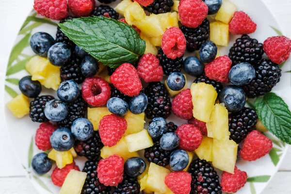 白いテーブルの上の皿に虹色やパイナップル ブラックベリー ブルーベリー ラズベリーとミントで健康的な果実のビューをクローズ アップ — ストック写真