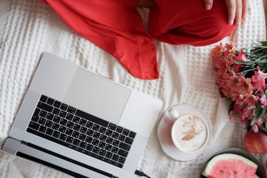Üstten görünüm kadın yatağın üstünde laptop üzerinde kırmızı bir elbise içinde elleri ve pencere arka plan karşı kahve içer 