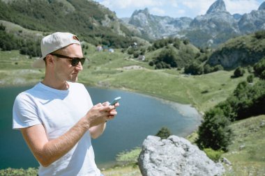 Gezgin bir kap ve güneş gözlüğü duran erkekte smartphone kullanarak ve güzel sahne dağ manzarası Karadağ görmek