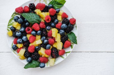 Gökkuşağı renkleri, ananas, böğürtlen, yaban mersini ve ahududu nane ile sağlıklı meyveleri üst bakış beyaz masada tabakta. Kopya alanı