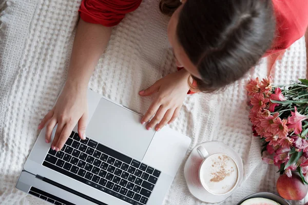 Top view of blonde woman in a red robe working on laptop on the bed and drinks coffee against window background