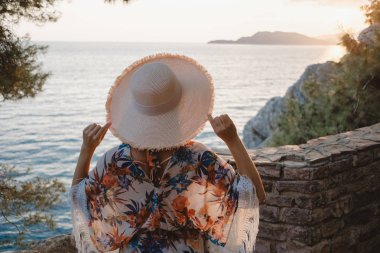Arkadan görünüşü plajda hasır şapkasını tutarak ve ufuk arıyorum genç güzel bir kadın.