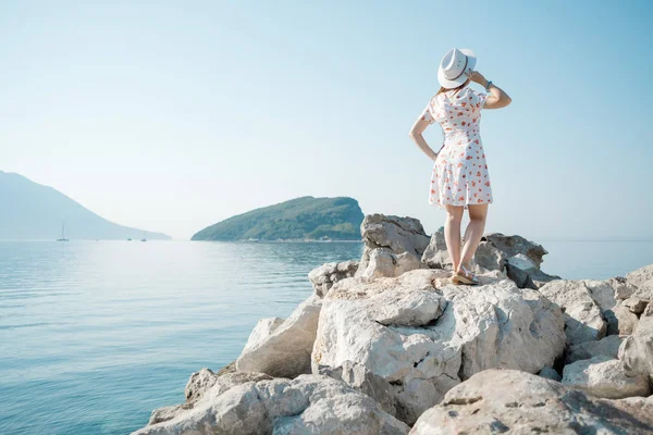 一个年轻美丽的女人在海滩上抱着她的白帽子的背面视图 并期待地平线 — 图库照片