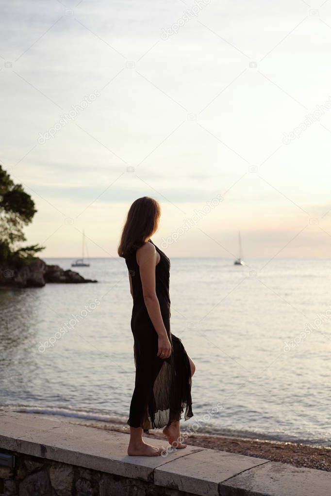 Back of a young woman in a black dress looking at the sea at sunset.