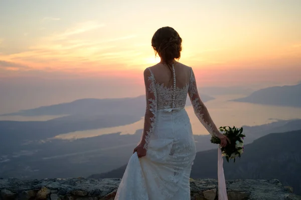 在日落时分站在山顶上的白色礼服的新娘的背 — 图库照片