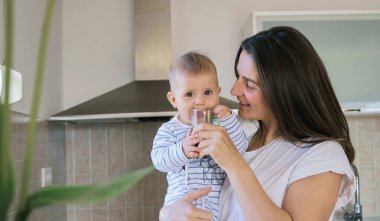 Beyaz tişörtlü genç bir kadın bebeğiyle oynuyor ve evde, mutfakta yeşil smoothie içiyor.