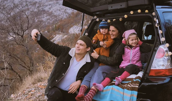 Family  sitting in the open trunk of a black car with kids and make a photo on the phone or selfie against the backdrop of autumn mountain