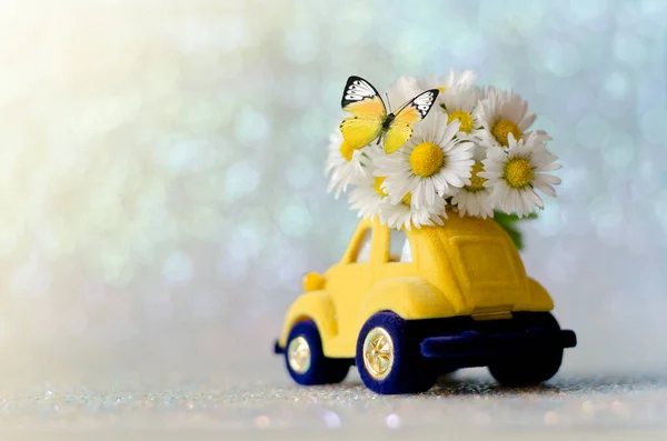 Card with a little toy car delivering white bouquet flowers on blue blurred background and butterfly.