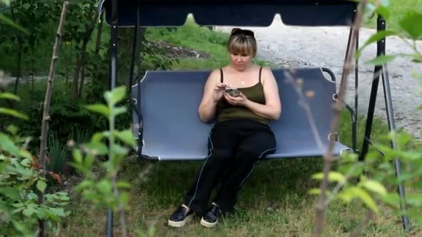 Молодая женщина сидит на качелях и смотрит на телефон — стоковое видео