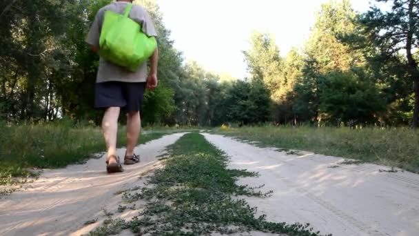 Um homem de calções com uma grande bolsa verde no ombro caminhando ao longo de uma estrada rural à distância — Vídeo de Stock