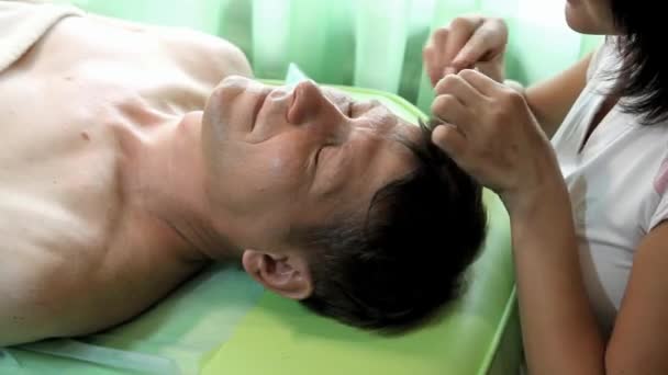 Kvinnliga kosmetolog avlägsnar hårstrån från ansiktet av en man som ligger på en grön stol — Stockvideo