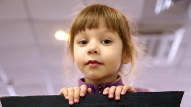 Little girl looks out from behind a chair and grimaces — Stock Video
