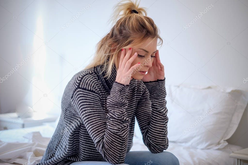 Sad young woman sitting on the bed at home suffering headache