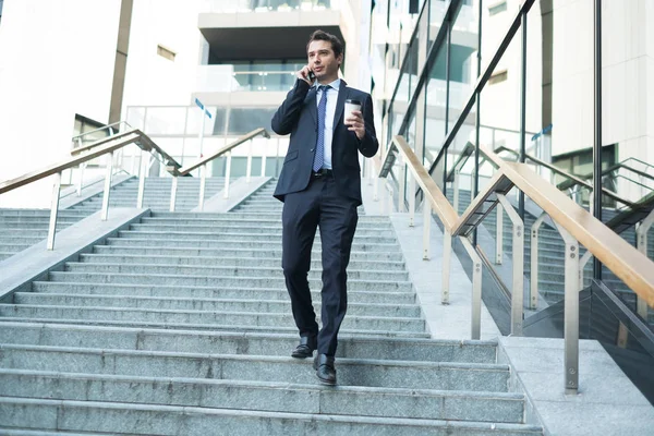 Confident manager with cellphone walking on stairs holding cup of coffee