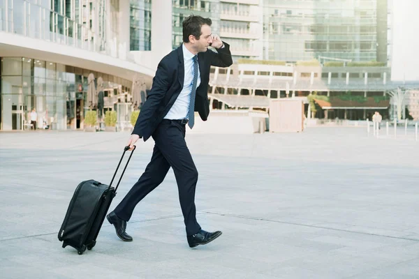 Stressed late businessman running with rolling bag and talking on the phone